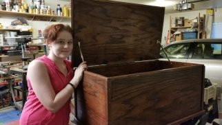 A young woman beside a large unfinished plain-wood chest, lid open
