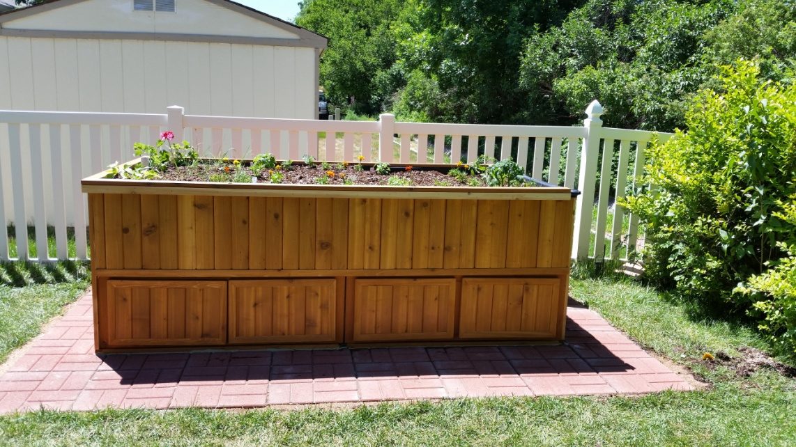 The elevated cedar garden bed, planted, on a brick pad by a white picket fence